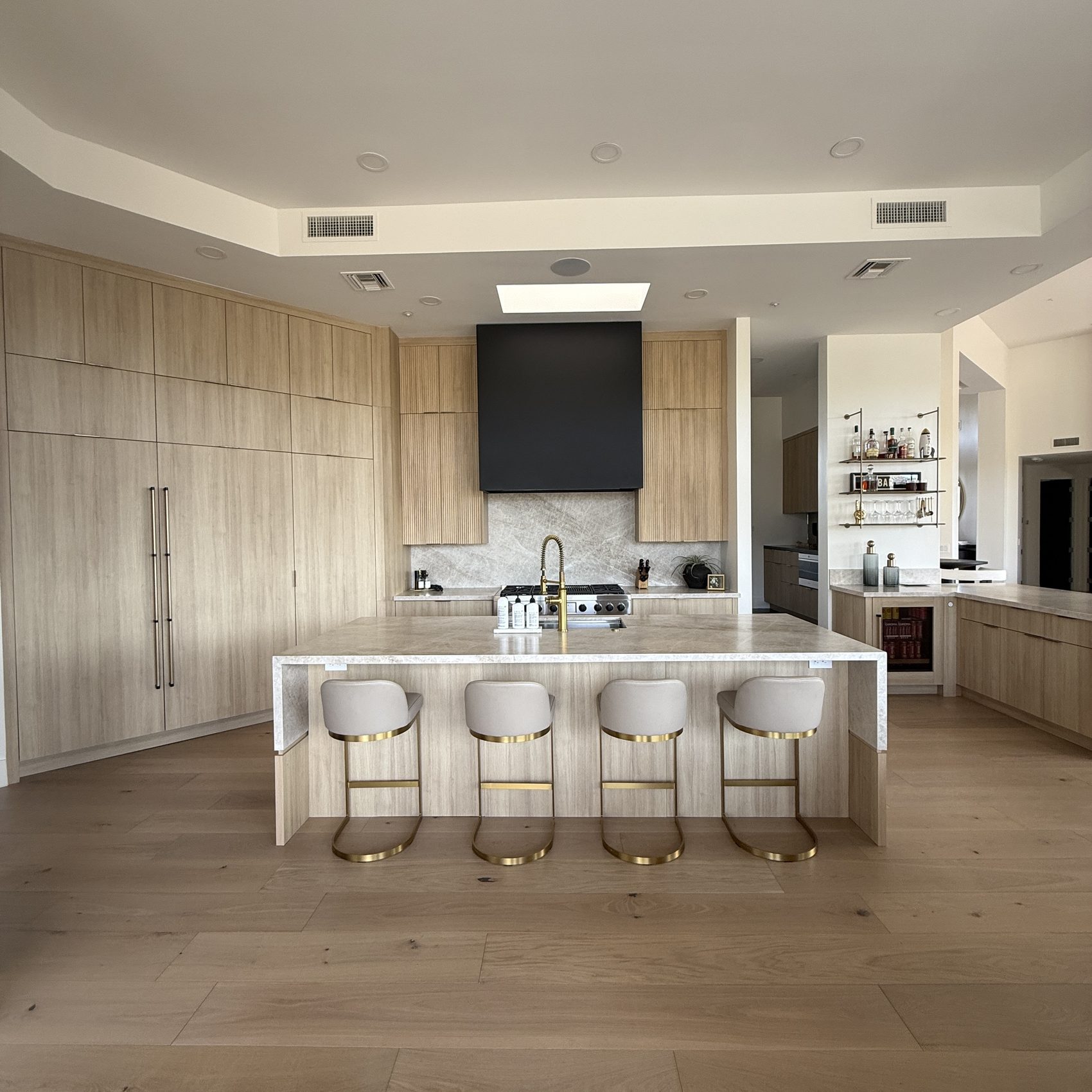 The same kitchen rebuilt — rift white oak, white quartz waterfall island, black hood and brass stools